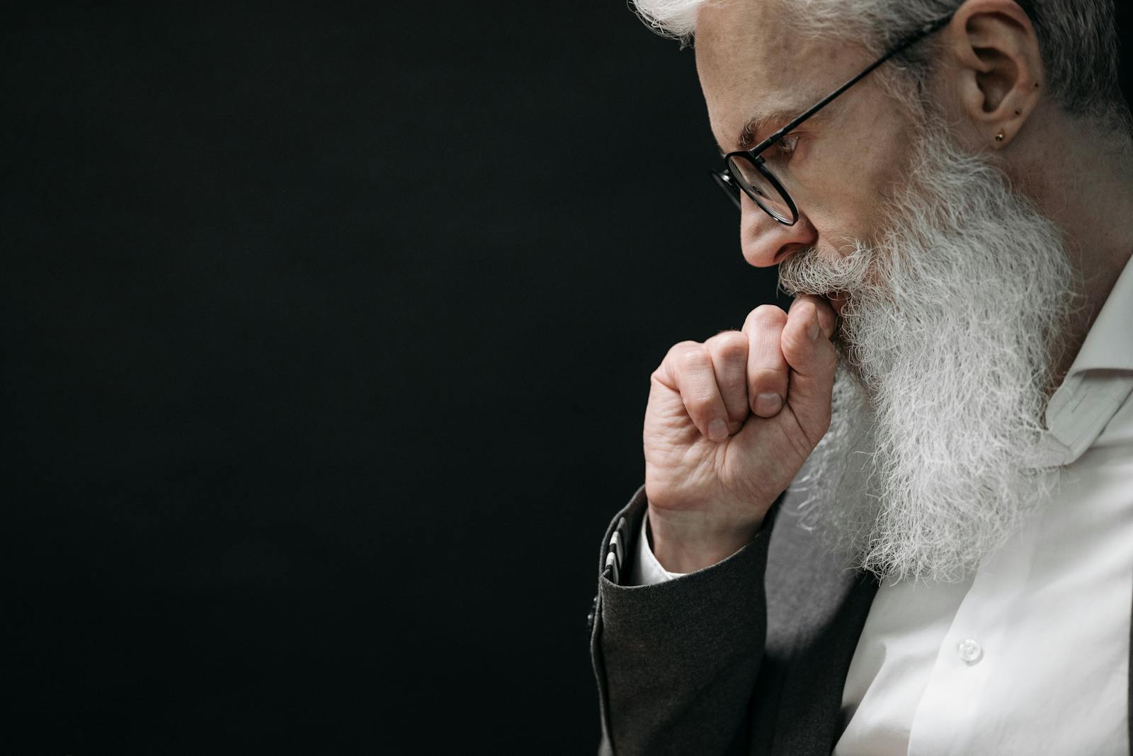 Close-up of an elderly man in deep thought, wearing eyeglasses with a dark background.